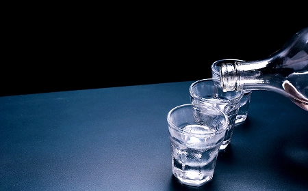 Прозрачная бутылка наливает крепкий алкоголь в три стопки на синей поверхности с чёрным фоном